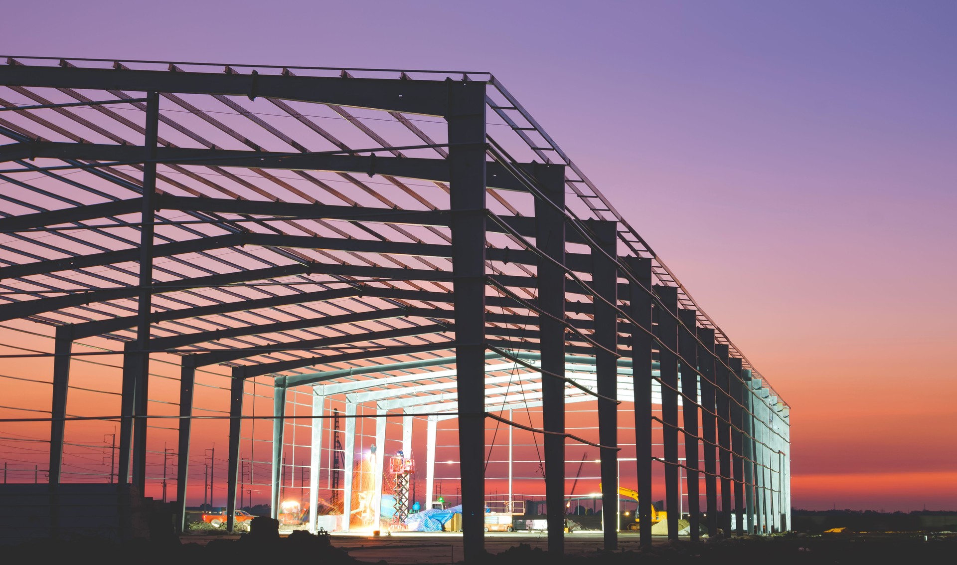 Silhouette of industrial factory building framework with welder workers working overtime to welding metal structure in construction site area against colorful twilight sky background